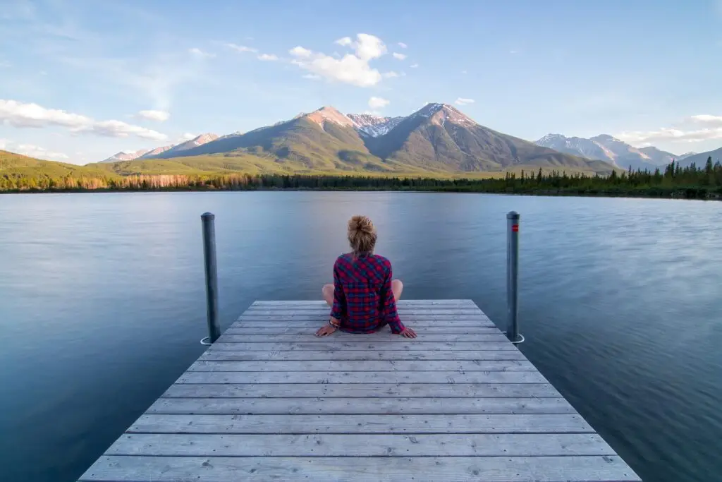 A girl sitting on a small jetty looking over a lake and mountains in the distance.